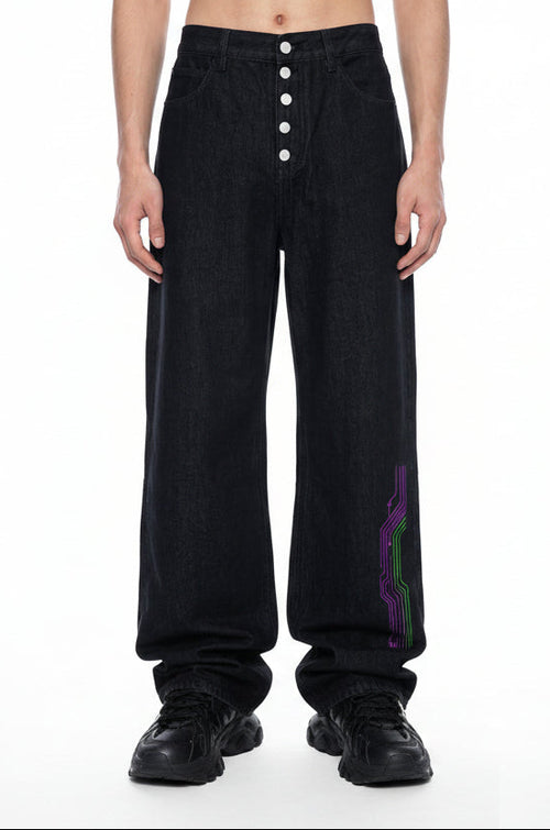 A model is shown from the waist down wearing black wide-leg denim pants with a button fly and a purple and green graphic detail on the lower left leg, paired with white socks against a white background.