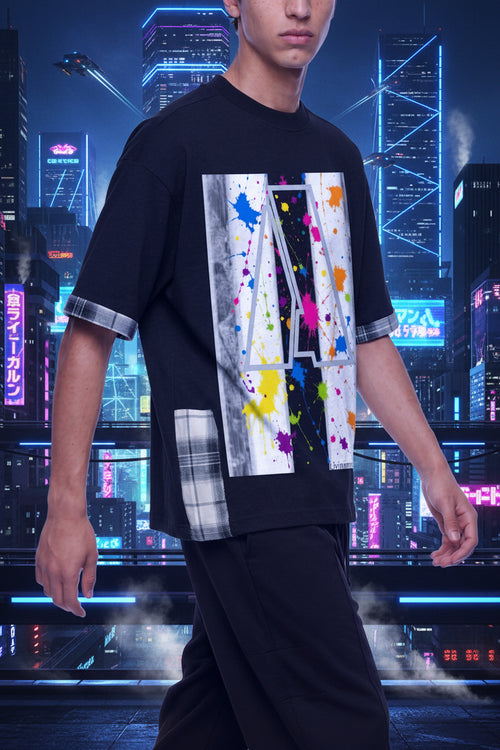 A man wearing a black short-sleeve t-shirt with a large abstract graphic print on the front featuring paint splatters in multiple colors, and plaid trim on the rolled-up sleeves and hem.