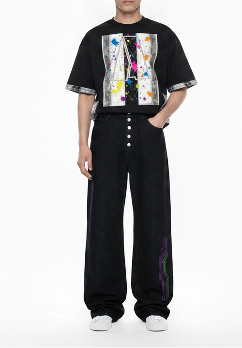 A model wearing a black graphic t-shirt with colorful paint splatters and striped sleeve cuffs, paired with black wide-leg button-front pants and white socks, against a plain white background.