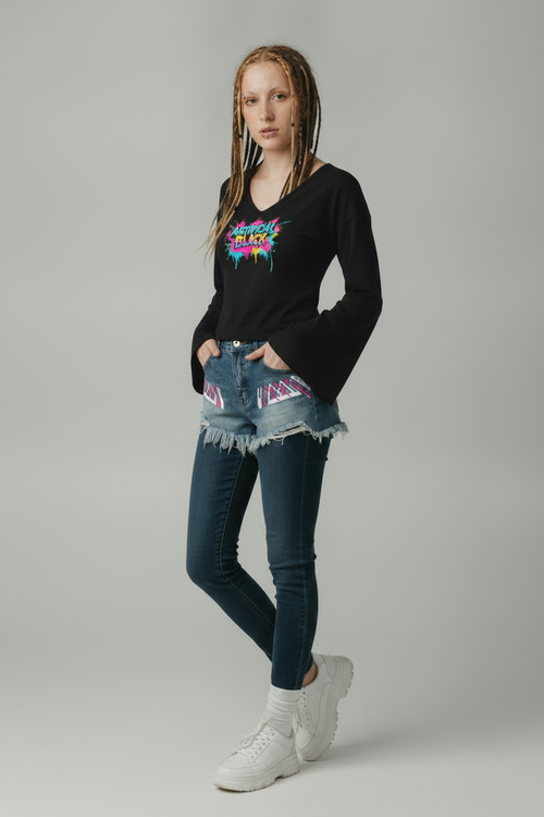 A model stands against a light gray studio background wearing a black long-sleeve graphic t-shirt, frayed denim shorts with colorful geometric patterns on the pockets, dark blue skinny jeans, white socks, and white sneakers.