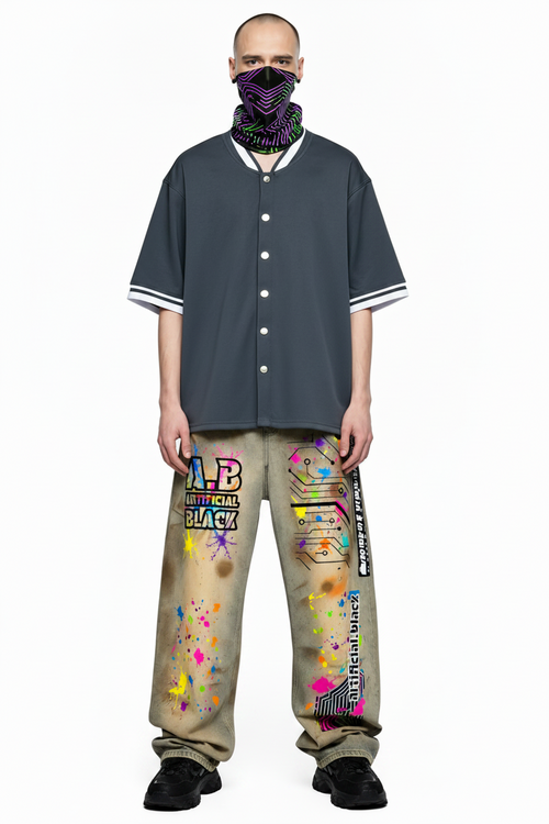 A model stands against a white background wearing a dark gray short-sleeve button-up shirt with white and black striped cuffs, beige pants covered in colorful graffiti-style graphics, and a black neck gaiter.